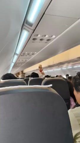 The video is a first-person view from inside an airplane cabin. The camera is pointed forward, showing the aisle and seats occupied by passengers. In the distance, a flight attendant in uniform is visible, standing and appearing to address the passengers or perform duties. The cabin interior is illuminated by overhead lights.