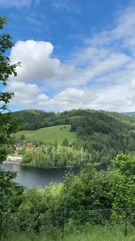 The video displays a slow panoramic pan of a scenic natural landscape in Wisła, Poland. A dark body of water, likely a lake or reservoir, occupies the midground, surrounded by rolling green hills and mountains. These hills are extensively covered with dense evergreen and deciduous trees, forming lush forests, with some patches of open green fields visible on the slopes. In the distance, several small buildings or houses are nestled among the greenery. The sky transitions from partly cloudy with noticeable blue patches on the left to a more overcast and grey sky on the right. The foreground features vibrant green bushes and trees, with a metal fence visible at the bottom of the frame. The video captures a serene and picturesque view of the mountainous terrain, accompanied by indistinct human speech in the background.