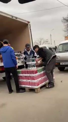 Three men are unloading cases of soft drinks from a truck's open cargo hold. The cases are stacked on a pallet, each containing several cans. The men are dressed casually in jackets and hats. The surrounding area appears to be an outdoor market or loading dock with other vehicles parked nearby. The sky is overcast. The cases on the pallet are primarily red, green, and black. The cans are stacked high and the men are carefully removing cases to distribute them. The truck is white and appears to be in an industrial or commercial setting.