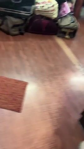 The video shows a pile of luggage and bags stacked against a wall. The luggage is of various sizes and colors, including a large black suitcase, a maroon duffel bag, and several smaller bags. There are also some pillows and a woven basket on top of the luggage. The floor is made of wood. The room appears to be a bedroom or storage area, with a window visible in the background. The lighting is dim.