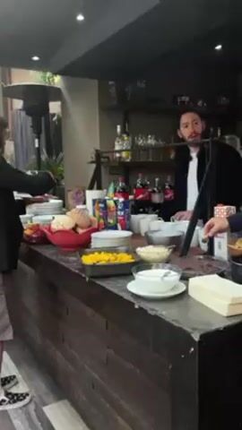 The video depicts a bar area indoors, likely during a brunch or similar event. There is a long counter with various food items displayed, including fruit, pastries, and sliced mangoes. Behind the counter is a man wearing a black jacket and white shirt, possibly a bartender or server. People are seen approaching the counter, seemingly helping themselves to the food. One person is wearing a black blazer, grey shorts and patterned shoes. The background includes shelves with bottles and glassware, suggesting it's a bar setting. A black outdoor heater can be seen through the back window.