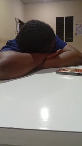 A person is sleeping on a white table with their head down and arms crossed. A phone is next to them on the table. There is a window with blinds and a calendar or sign on the wall behind the person. The room appears to be indoors with yellowish lighting.