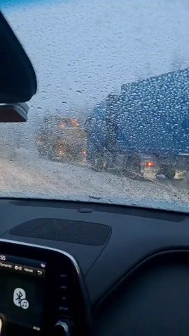 The video, shot from inside a car, shows a snowy day with poor visibility. The windshield is covered in raindrops and snow. A large blue truck is visible in front of the car. Inside the car, the dashboard is visible. A phone is attached to the dashboard displaying a navigation app, and the car's entertainment system is showing music information. A string of beads hangs from the rearview mirror.