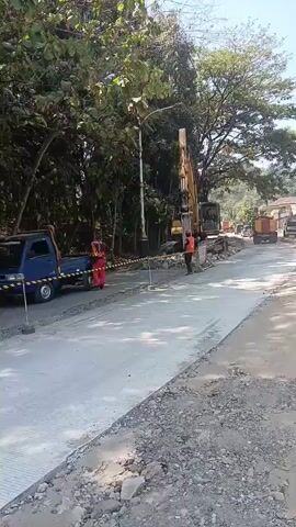 A construction site is shown, with a road being repaired or built. A yellow excavator is visible, as are two trucks, one blue and one red and yellow. Two individuals in orange vests are near the excavator, and one appears to be operating it. Two people are riding a motorcycle, and another person is standing near the blue truck. The area is surrounded by trees and a light pole is also visible. The road surface is a mixture of gravel and newly laid concrete.