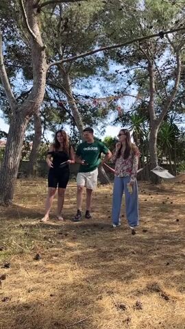 Three people stand in a grassy area with scattered pine cones, beneath tall trees. The middle person, a male, wears a green Adidas t-shirt and white shorts, and checks his phone. The woman on the left wears a black dress, while the woman on the right wears a floral top and blue slacks. Each person holds a yellow object, potentially a piece of paper. The woman on the right raises her arms and cheers, the other two join in, and the woman on the left giggles. A hammock hangs in the background, adding to the relaxed atmosphere of the scene.