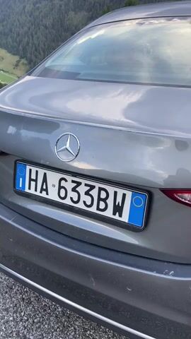 This video showcases the rear of a gray Mercedes-Benz sedan parked on a gravel road overlooking a breathtaking mountain valley in Ratschings - Racines, Italy. The license plate reads "HA 633BW," indicating it is an Italian vehicle. The car is positioned on a scenic overlook, with a guardrail running along the edge of the road.

The scene is set in a mountainous region characterized by steep, tree-covered slopes and a winding valley below. Lush green meadows and dense forests dominate the landscape, with a small village nestled in the distance. The sky is a dramatic mix of blue with scattered, heavy clouds, suggesting either approaching or departing rain, and the lighting indicates it is likely late afternoon or early evening. The overall impression is one of serene beauty and the vastness of nature, with the car serving as a point of human presence within this grand setting. There are no people visible in the frame.