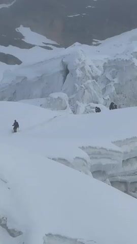 The video pans across a snowy, mountainous landscape with large ice formations. In the distance, a group of people dressed in winter clothing can be seen walking and standing on the snow. Further in the foreground, several tents are set up in the snow. The sky is overcast and foggy, creating a muted, monochrome scene. One tent is red with a yellow logo that appears to be "PIONEER CAMP." Another tent is teal blue. Two people are standing near the red tent, and their gear is visible. The ground is covered in deep snow, with some areas showing the texture of ice. The overall impression is of a harsh, cold environment, likely a high-altitude expedition camp.