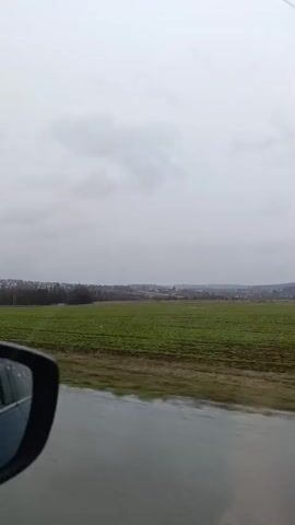A vertical shot from a car moving on a road shows a landscape outside. The sky is overcast and a field of green stretches to the treeline in the distance, where buildings are visible on a small rise. Rain streaks the road. The side mirror of the car shows a reflection of a person looking at a cell phone inside the car.