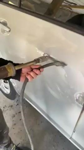 A close-up shot depicts a car repair process on a silver car door. A person holds a yellow-colored tool with a nozzle and a black hose attached, which is used to apply heat to a specific area of the door. The door surface appears to have been treated with a white substance, possibly filler or primer, and there's a distinct dent being worked on. A tire can be seen in the periphery of the shot, providing context to the automotive setting. The overall lighting is bright, highlighting the details of the repair work.