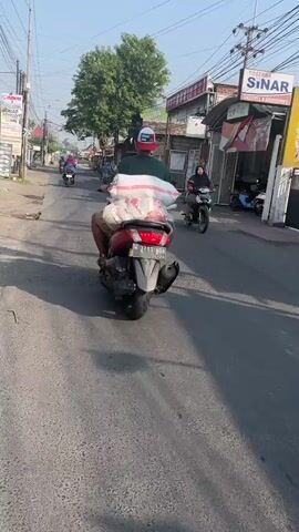 A motorcycle rider, wearing a helmet and a white and green shirt, carries a large bundle of white sacks on the back of their red scooter. They are riding down a paved road with other motorcycles and buildings lining the sides. The sky is clear and the sun is shining.