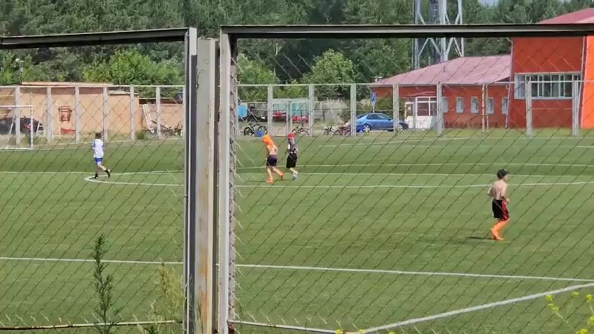 The video shows a soccer field with three young boys playing. Two boys are in the foreground, and one is further back. The boy on the left is wearing a white shirt and blue shorts. The boy in the middle is wearing an orange shirt and dark shorts. The boy on the right is wearing an orange shirt and orange shorts and a red hat. The field is green with white markings. In the background, there is a fence, some buildings, trees, and a car. The weather appears to be sunny. The video is filmed through a chain-link fence, which is visible in the foreground.