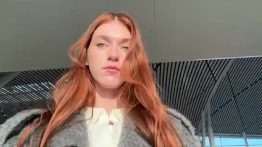 The video begins with a close-up shot of a woman with long, red hair, wearing a gray, textured coat over a white shirt. Her gaze is directed towards the camera. The camera then pans to the right, revealing the interior of an airport terminal. Seating areas with people are visible through the large windows, which offer a view of the outside. The camera angle shifts to showcase the airport runway bathed in sunlight. Several airplanes are parked at the gates, with airport vehicles and personnel visible on the tarmac. In the background, distant mountains can be seen. The overall atmosphere suggests a bright, sunny morning at an airport.