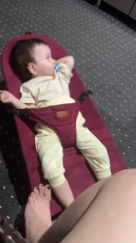 A baby lies in a maroon baby bouncer with black straps. The baby is wearing a light-colored t-shirt and light yellow pants with a pattern. The baby has a pacifier in its mouth and is resting its right hand on its chest. The baby bouncer is on a grey carpet with a dotted pattern. In the foreground, a person's legs and feet are visible, with the left foot being closest to the baby bouncer. The person's left foot has black nail polish.