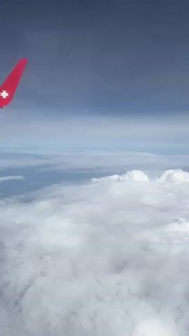 From the vantage point of an airplane window, a view is captured showing the plane's red wing against a backdrop of clouds and sky. The clouds form a dense, white layer, resembling a sea of cotton. Beyond the clouds, a strip of light blue sky is visible, meeting a darker blue-gray expanse higher up. The plane's wing, situated on the left side of the frame, features a white star-like emblem.