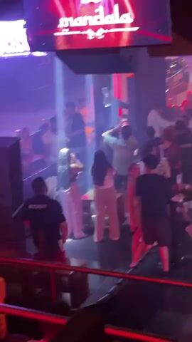The video shows a crowded nightclub, with people dancing and socializing. The lighting is dark, with colored lights and spotlights illuminating the scene. There are several people visible, of varying ages and ethnicities. Most are dressed casually. Some are drinking and appear to be enjoying themselves. A red decorative lantern sits on a pole in the middle of the room. A security guard is seated and watching over the crowd. A sign that says "Mandala" is visible above. The music is loud and upbeat, typical of a nightclub setting.