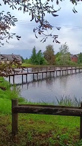 A wooden bridge extends across a body of water. On the far bank, there are trees, houses, and a gazebo. The foreground shows grass and a wooden fence. The sky is overcast.