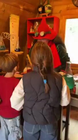 Inside a wood-paneled room, a person dressed as a whimsical wizard stands behind a counter, engaging with two children. The "wizard" wears a red pointed hat with goggles, a green jacket, and a red scarf. The children, a boy in a red sweater and a girl in a black vest, stand facing the wizard, seemingly captivated. Various props, including glass beakers filled with colored liquids, a red shelving unit with small bottles, and decorative plants hanging from the ceiling, contribute to the fantasy atmosphere. The wizard appears to be stirring a concoction with a wooden stick.