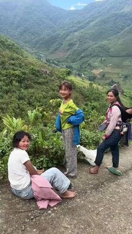 The video shows a mountain path in a rural setting. Two young women are standing on the path. One woman has a baby strapped to her back.  A woman wearing a sun hat and carrying a backpack is walking down the path away from the camera. In the background, mountains covered with green vegetation can be seen. There are terraced fields in the distance. The sky is partly cloudy.