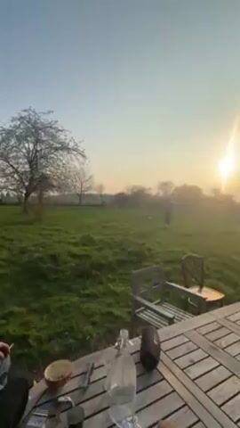 The video shows a sunset over a grassy field. A wooden table and chairs are in the foreground. There are several people in the distance, seemingly playing. A bottle and a speaker sit on the table.