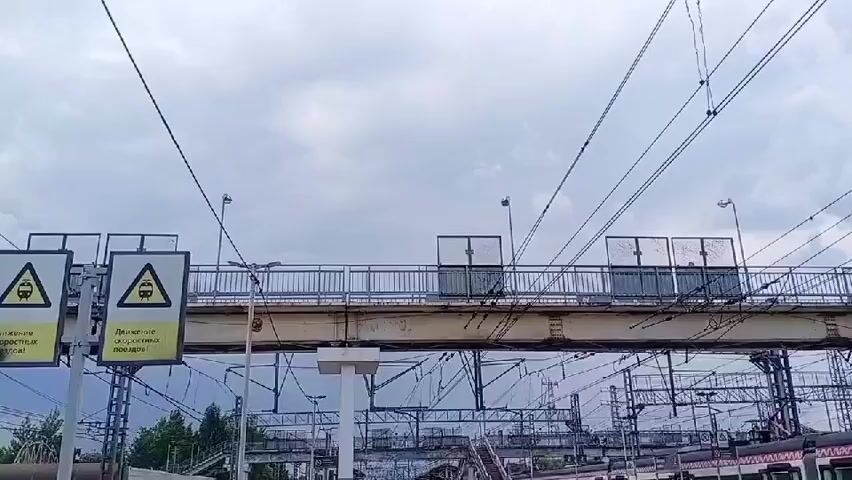 This media file captures a view of a railway overpass on an overcast day in Fryazevo, Russia. The overpass is a prominent object, featuring a metal railing and several transparent enclosures that appear to be shelters or viewing areas. Below the overpass, a complex network of railway tracks and overhead power lines for electric trains is visible.

Two yellow warning signs with black triangular borders are prominently displayed on the left side of the frame. Each sign depicts a black silhouette of a train. The text on the left sign reads "Движение скоростных поездов!" which translates to "Movement of high-speed trains!". The text on the right sign is partially obscured but appears to be the same.

The sky is filled with heavy, grey clouds, indicating a cloudy or possibly rainy day. The time of day is likely daytime, given the lighting conditions, but the overcast sky diffuses the light. The scene is a functional railway environment, suggesting a transportation hub or a busy route for trains. There are no discernible people in the foreground or midground, though there might be figures within the transparent enclosures on the overpass, too indistinct to identify. The overall mood is utilitarian, focusing on the infrastructure of the railway system under a subdued sky.