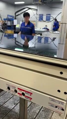 The video shows a factory setting where insulated glass units are being produced. A man in a blue uniform is working on a glass unit, which is lying horizontally on a metal table. He seems to be sealing the edges of the glass. The glass is reflective, mirroring the worker. In the background, there are machines and other workers. There are two women walk by, one of whom is looking at her phone. Another man follows the women.