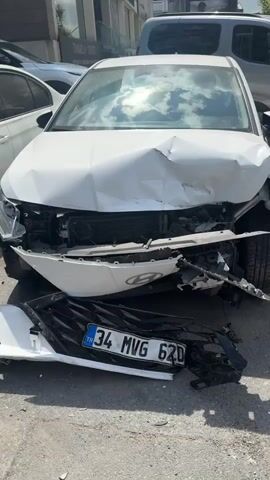 A white Hyundai car is depicted with significant front-end damage. The hood is heavily dented and crumpled inwards, obscuring the radiator and engine components. The front bumper is detached and lying on the ground in front of the car, along with pieces of the black grille. The license plate, which reads "34 MVG 62D" and appears to be Turkish, is also dislodged and on the ground. The car's headlights are damaged and partially obscured. The vehicle is parked on a paved surface, likely a street or parking lot, with other vehicles and buildings visible in the background. Sunlight is reflecting off the car's windshield and hood, indicating it is daytime. There are no people visible in the video.