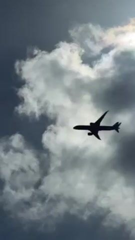 A passenger airplane is flying overhead. It starts silhouetted against a cloudy sky and then transitions to a clear blue sky as the video progresses. The plane is white with green and blue markings.