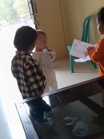 A video clip shows two young children in a room. The child on the left is wearing a plaid shirt and dark pants and is looking towards a doorway. The child on the right is wearing a striped shirt and light pants and is holding a piece of paper. A green plastic chair is visible in the background. The children appear to be interacting, though their specific actions are unclear due to the low resolution of the video.