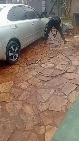 A person is pressure washing a silver sedan parked on a flagstone driveway. The person is bent over and using a pressure washer on the car's tires and lower body. Water is spraying from the pressure washer, and the driveway is wet. There is a green hose connected to the pressure washer. Other objects visible include a small generator, a bucket, and a fence.