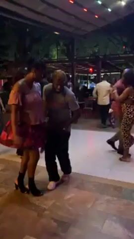 The video shows a group of people dancing together outdoors, in what appears to be a social gathering. The setting is under a covered structure, with string lights overhead adding to the ambiance. The floor has a patterned tile design. A couple in the foreground is the main focus: a woman in a blush-colored off-the-shoulder top and a floral skirt, and a man in a gray t-shirt, dark pants, and a beige baseball cap. They are dancing closely together, moving rhythmically to music. Other couples are also dancing nearby, including one woman wearing a leopard-print dress. Trees and foliage can be seen in the background, suggesting a garden or park setting. The lighting is soft, creating a relaxed and joyful atmosphere.