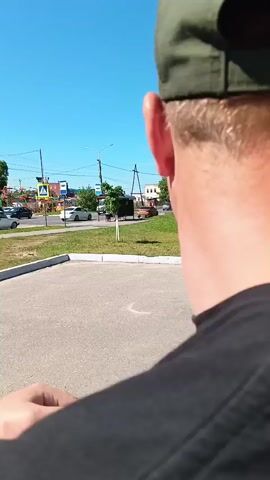 A person films from over their shoulder as a line of people walks toward a yellow bus and gets on. Another person with a backpack gets out of a yellow van, and people are around another yellow bus. In the foreground, the top of a white car with a blue light on its roof is visible. The scene takes place outdoors on a sunny day.