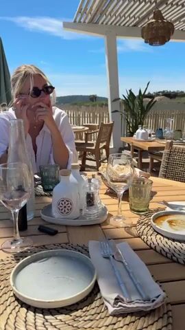 The video showcases an outdoor dining experience at a beachside restaurant. A woman wearing a white shirt and sunglasses sits at a wooden table set with tableware, including wine glasses, plates, and condiments. The table is adorned with woven placemats. The background reveals a sandy beach, the ocean, and a clear blue sky. Beach huts and umbrellas are visible along the shoreline. Palm trees add to the tropical ambiance. The camera pans around the scene, emphasizing the proximity of the restaurant to the beach.