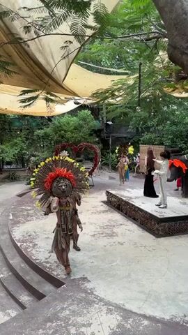 The video shows a procession of people in elaborate costumes, seemingly for a performance or parade. In the foreground, a person wearing a large feathered headdress with a lion's face motif and tribal-inspired clothing walks towards the camera. To the right, a raised platform is visible, with several individuals dressed in bright, revealing outfits, some adorned with feathers, walking along it. In the background, more participants in colorful costumes are seen, and a large heart-shaped floral decoration is present. The environment appears to be outdoors, with lush greenery and a covered canopy overhead, suggesting a tropical or garden setting. The overall atmosphere is festive and theatrical.