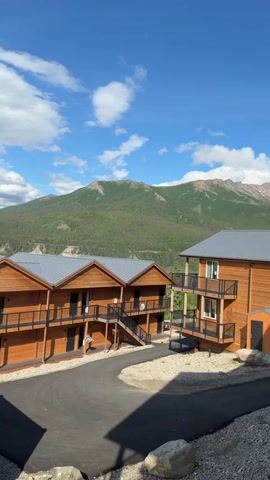This media file depicts a scenic view of buildings nestled against a backdrop of majestic mountains and lush green forests. The buildings are constructed with warm-toned wood, featuring multi-story designs with balconies and metal roofs. A paved driveway curves through the property, surrounded by gravel and natural landscaping.

The dominant subjects are the architectural structures and the vast natural landscape. The setting is clearly outdoors, with a strong emphasis on the natural beauty of mountains and forests. There are no people visible in the media. The activity taking place is purely observational, capturing a tranquil moment of the landscape and its architecture.

Notable details include the time of day, which appears to be late afternoon or early evening, given the quality of light and the presence of shadows. The weather is fair with a mix of blue sky and fluffy white clouds, suggesting pleasant conditions. The location cues, such as the dense evergreen forests and snow-capped mountains in the distance, strongly suggest a mountainous region, consistent with the provided context of Denali National Park and Preserve. The overall emotion evoked is one of peace, serenity, and awe at the natural grandeur. There is no visible text in the media file.