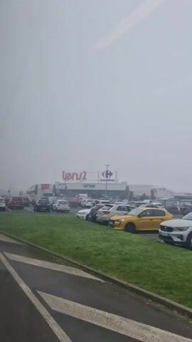 The video shows a partially full parking lot on a foggy day outside of a Carrefour supermarket, with associated stores, including ACTION and Lunch, visible on the side of the building.  The parking lot is relatively full with a variety of cars parked in the spaces.  There is a grassy area that is adjacent to the parking lot with trees visible in the distance behind the building.  The sky is overcast and the weather is foggy, reducing visibility.  The camera is moving on a vehicle.
