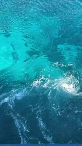 The video shows a top-down view of clear turquoise water with darker blue water in the foreground. The lighter turquoise water appears shallow and possibly has sand or seabed features visible beneath the surface. In the center of the frame, there is a disturbance in the water, creating white foam and swirling patterns. This disturbance suggests movement or activity, possibly a jump or dive into the water, or a natural phenomenon like a current or undertow. There are no people or objects directly visible in the frame. The environment is clearly a body of water, likely the ocean or a very large lake, given the depth variation and color.