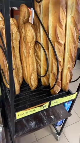 This is a close-up, eye-level video of a display of freshly baked baguettes in a store. The baguettes are golden brown with distinct scoring marks on top. They are arranged on a black metal rack. Yellow price tags are visible, one labeled "TRADITION 1€20" and another labeled "BAGUETTE 1€10". The background is slightly blurred but shows shelves with various products, suggesting a grocery store or bakery. The lighting is bright, highlighting the texture and color of the bread. There are no people visible in the frame. The video pans slightly to showcase the different baguettes and their pricing.
