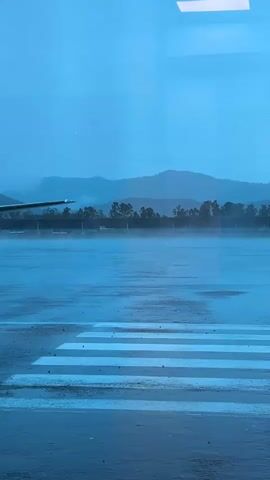The video shows a wet airport tarmac under heavy rain, viewed through a window with a blue tint. In the background, misty mountains are visible. A white and blue twin-engine propeller airplane with landing gear extended slowly taxis from the right side of the frame towards the left. The tarmac surface is highly reflective due to the standing water, and white pedestrian or ground markings are visible in the foreground. A rectangular light fixture is reflected in the top right corner of the window.