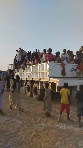 Here's a description of the video:

The video shows a large group of people riding on the back of a flatbed truck. The truck bed is crowded with people, mostly children and young adults. Some people are sitting, while others are standing. Several people are also standing around the truck, and walking away from it. The people are wearing a variety of casual clothing. The truck is parked on a dirt road or open area, with a clear sky in the background.  The atmosphere appears lively, with music playing in the background.