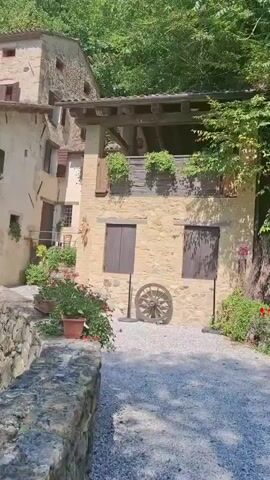 The video pans across a rural Italian village scene. The buildings are constructed of stone with wooden shutters and balconies adorned with plants. A water wheel is prominently featured, situated next to a body of water. The surrounding environment is lush and green, with trees and vegetation covering the hillsides. A small waterfall cascades down a rocky cliff face into the water. The overall impression is one of rustic charm and natural beauty.