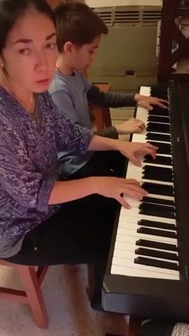 The video shows two people, a woman and a boy, playing a digital piano. They are sitting side-by-side on red stools, facing the piano. The woman is on the left, and the boy is on the right. They are both looking at the piano keys as they play. The woman is wearing a purple patterned shirt, and the boy is wearing a blue shirt. The piano is black and appears to be a full-sized keyboard. The background is blurry, but it appears to be an indoor setting with a wall and a window visible. The lighting is dim. The video is shaky and appears to be handheld. The audio contains the sound of the piano playing.
