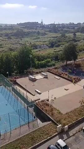 The video shows an aerial view of a park with a skatepark, a playground, and a tennis court. There are also several cars parked along a street in front of apartment buildings. The buildings are mostly residential, with some commercial spaces on the ground floors. The park is well-maintained, with trees and landscaping. The weather appears to be sunny and clear. There are no people visible in the video.