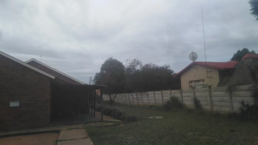 The video begins with an upside-down view of a cloudy sky, a house with a red roof, a fence, trees, and a brick building. The camera then moves quickly, showing a person’s leg in blue jeans and a white sock. The background is a blurry light brown surface.