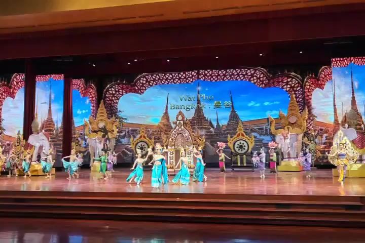 A group of dancers in blue and gold traditional attire perform on a stage in front of a large LED screen. The screen displays a vibrant backdrop of ancient Thai temples and pagodas, with ornate golden structures and statues of elephants on either side. The dancers move gracefully in unison, their movements synchronized and fluid. Some dancers hold colorful fans, while others gesture with their hands and arms. The stage is well-lit, highlighting the performers and the detailed backdrop. The overall scene suggests a cultural performance or theatrical production, likely depicting a scene from Thai history or mythology.