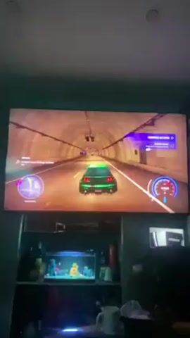 The video shows a person playing a racing video game on a large flatscreen TV. The game features a green car driving through a tunnel. The scene then transitions to a city street. Below the TV is a shelf unit containing a small aquarium, along with other miscellaneous items. The TV is mounted on a wall in a room with a white ceiling.
