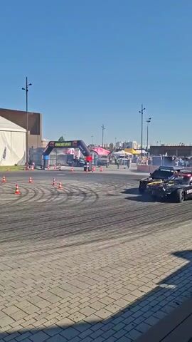 A wide shot captures an outdoor car drifting event under a clear blue sky. In the foreground, a gray paved area shows tire marks from previous drifts. In the middle ground, several cars are actively drifting, producing visible smoke. One car in the foreground, black with yellow and blue lettering that reads "INSTRUIRE AUTO", is turning right. Another car, dark purple, is drifting in the center. Further back, a blue car is also drifting. Behind the cars, an inflatable archway with sponsors' logos spans the track. To the left, a large white tent structure with a white banner is visible. The background shows more tents, vehicles, and trees, suggesting a festival or event setting. Orange cones are placed as markers on the track. The overall atmosphere is dynamic and energetic, focused on the motorsport activity.