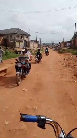 The video begins with a first-person perspective of someone riding a motorcycle down a dirt road. In the foreground, the handlebars of the motorcycle are visible. Several other motorcycles, each carrying multiple passengers, are traveling in the same direction, ahead of the viewer. The road is unpaved and reddish-brown in color. On either side of the road are buildings, some appearing to be houses or small shops, with fences and vegetation. Utility poles with electrical wires are visible along the roadside. The sky is overcast, suggesting a cloudy day. The overall impression is a rural or semi-urban environment, likely in a warm climate, where motorcycles are a common mode of transportation. The people on the motorcycles are dressed casually, and their faces are partially obscured. There is no discernible conversation or sound in the video, only the visual depiction of movement and the environment.