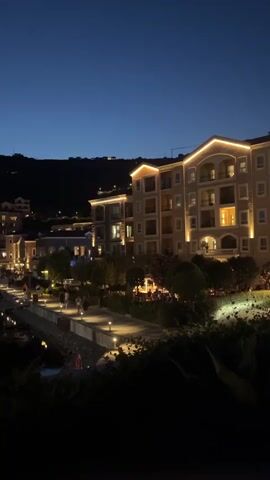 The video pans across a marina at dusk. Numerous boats are docked, and the buildings along the waterfront are illuminated with warm, inviting lights. The sky is a deep blue, and a few distant lights dot the hillside. People are seen walking along the promenade, enjoying the evening. The scene conveys a sense of tranquility and leisure.