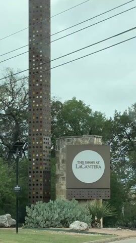 The video shows a car driving towards “The Shops at La Cantera” entrance. The camera shows the sign for the shopping center which has a stone base and a square sign with a white circle and the name of the shopping center. Next to the sign is a tall colorful pillar with many colored circles in a grid-like pattern. The road is asphalt and lined with trimmed hedges and small landscaping plants and rocks. The trees are green and full. The sky is overcast. The audio includes what seems to be a GPS system giving directions.