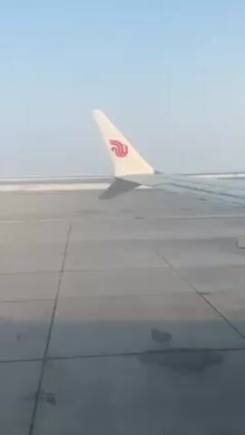 The video shows an airplane wing with the logo of Air China on the winglet. The plane is on a concrete taxiway or runway. In the distance, there appears to be a flat, white surface, possibly covered in snow or ice. The sky is a light blue. There are no visible people. The camera appears to be inside the plane, looking out the window.